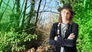 Craig Jordan-Baker image description: Craig is a white, middle-aged man with shoulder-length brown hair, wearing a dark hat and jacket. He stands in front of a forest scene.