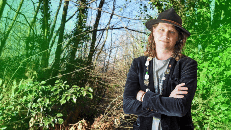 Craig Jordan-Baker image description: Craig is a white, middle-aged man with shoulder-length brown hair, wearing a dark hat and jacket. He stands in front of a forest scene.