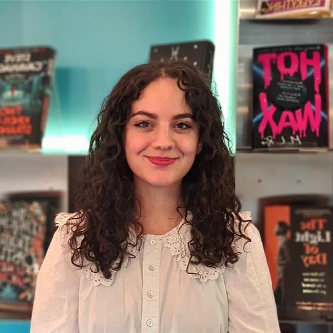 Jessie Goetzinger, a young white woman with dark curly hair and a white top. Books behind.