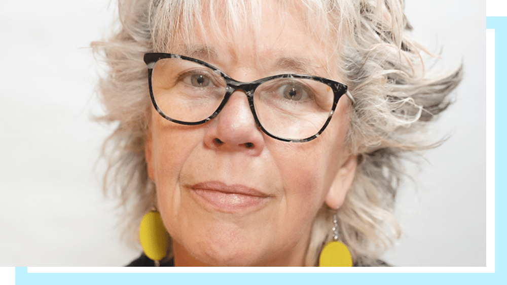 Sam Davey, a middle aged white woman with styled mid-length grey hair, glasses and yellow earrings