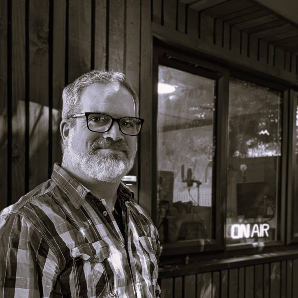 Simon Patrick, a middle aged white man with a beard and glasses stands in front of a radio studio