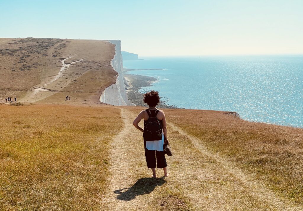 The writer Alinah Azadeh stands on the cliffside path approaching Birling Gap. The grass is yellow from summer. The sea is blue. A few people are in the far-off distance.
